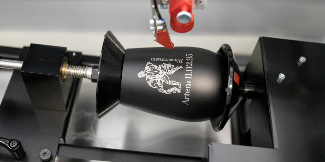 Trotec laser engraving machine marking a black metal tumbler with detailed artwork and text