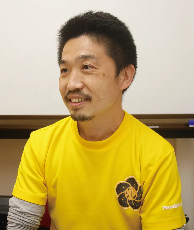 Man in yellow shirt with flower logo, seated indoors and smiling