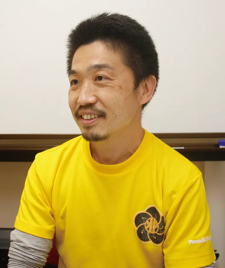 Man in yellow shirt with flower logo, seated indoors and smiling
