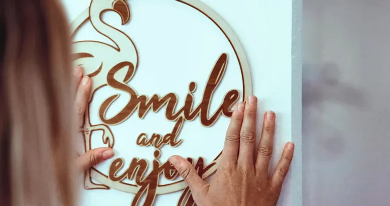 Hands installing laser-cut wood sign reading 'Smile and Enjoy' made with Trotec laser cutter