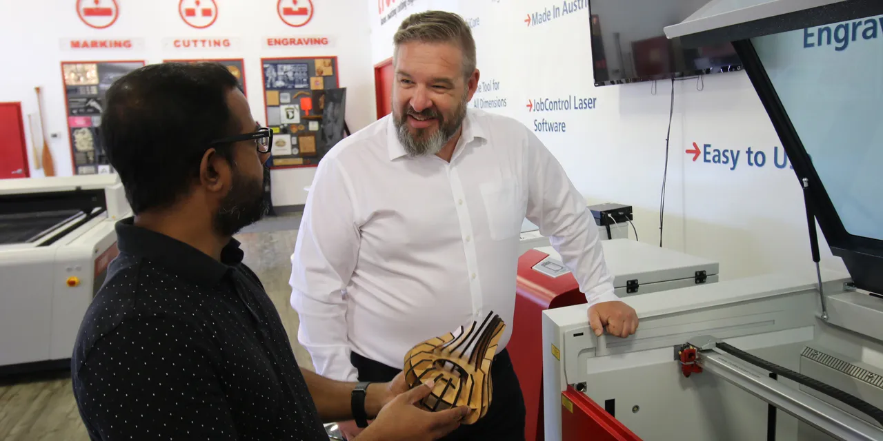 Two men discussing wooden model beside Trotec Speedy laser cutter in demo room